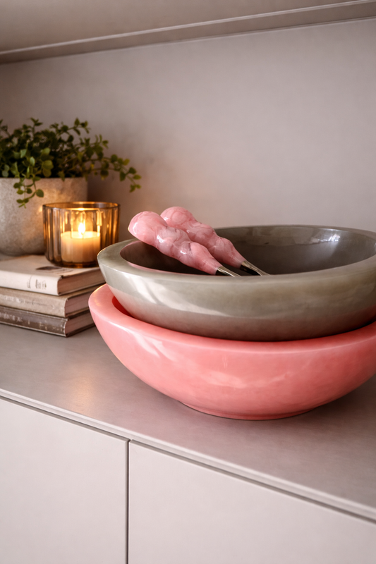 Two Ohana bowls, one shell pink and one pebble, on a surface with candles and books in the background.